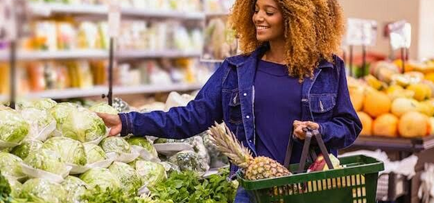 Woman with curly hair shopping in a market holding a green basket with pineapples and vegetables