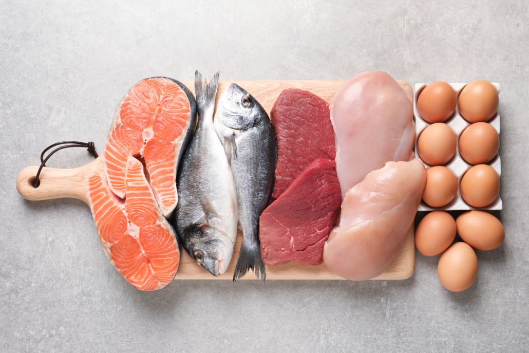 Cutting board with salmon slices, whole fish, raw beef, raw chicken breast, and eggs arranged on a gray surface